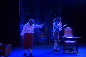 Scène de Voltaire aux Délices à Ferney-Voltaire : sous une lumière bleue, un homme aux cheveux gris, en chemise à jabot et culotte de brocart rouge, tend le bras ; face à lui, un jeune homme en gilet rayé lit une lettre.